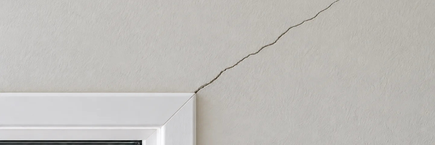 A diagonal crack running up from the corner of a white window frame on a light gray wall
