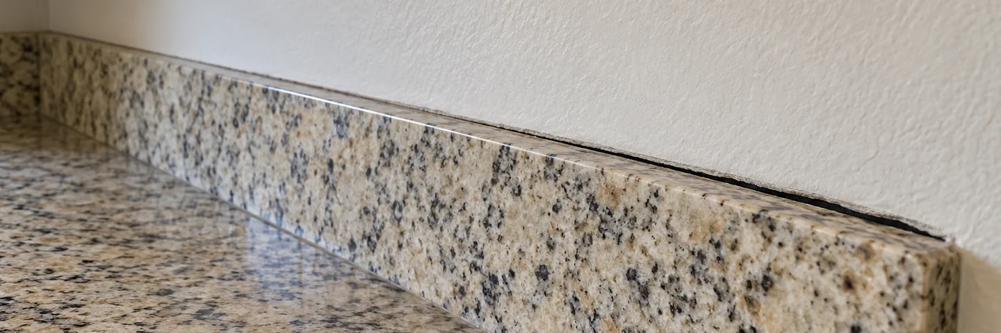 A thin gap opening between a granite backsplash and a textured wall under kitchen cabinets